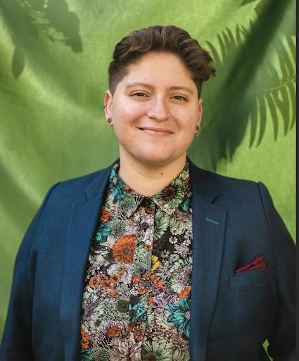 A photograph of a white queer person with short brown hair in a dapper navy suit jacket, a floral shirt, and maroon pocket square. They are smiling in front of a flowy green backdrop with shadows of plants. Headshot by Andrea Arevalo.
