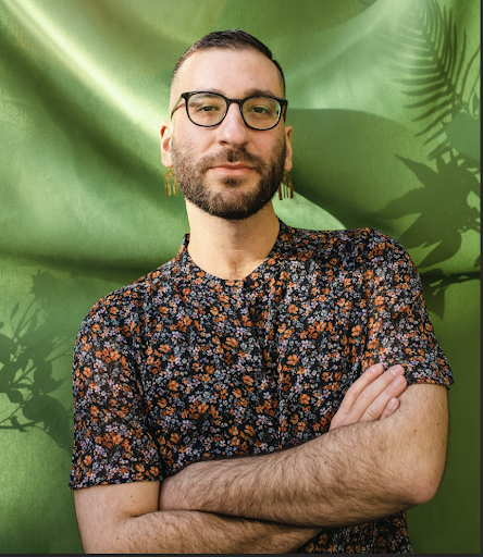 A queer, genderqueer, Arab artist and activist with glasses, a trim beard and a crew cut. They are wearing a floral top and gold dangle earrings. They are standing confidently in front of a flowy green backdrop with shadows of plants. Headshot by Andrea Arevalo.