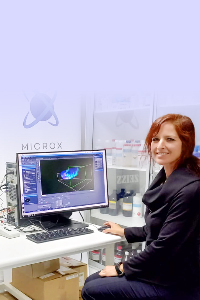 Alenka Lovy smiling in a lab, posing in front of a computer with imaging software open next to a light-sheet microscope.