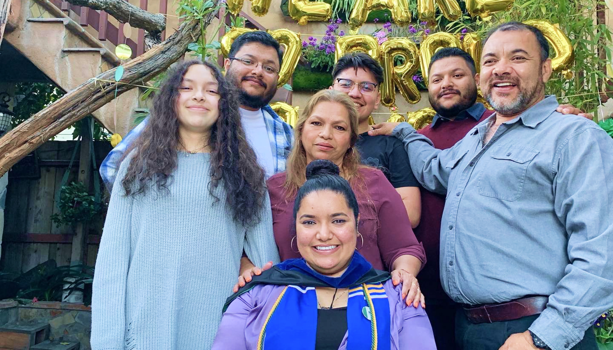 A group of people pose for a photo. In the center front is Mirna Cervantes, wearing graduation garb.