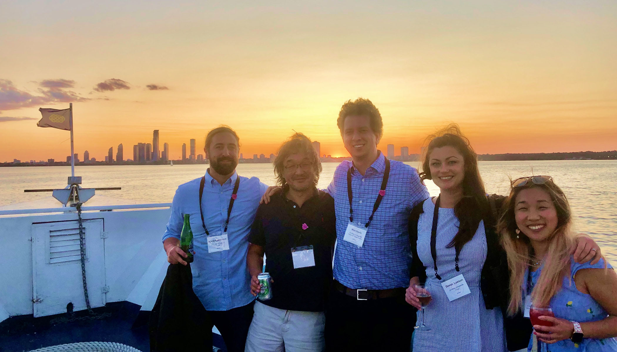 Five researchers smiling with arms around each other’s back while on a boat with the sunset and city skyline in the background.
