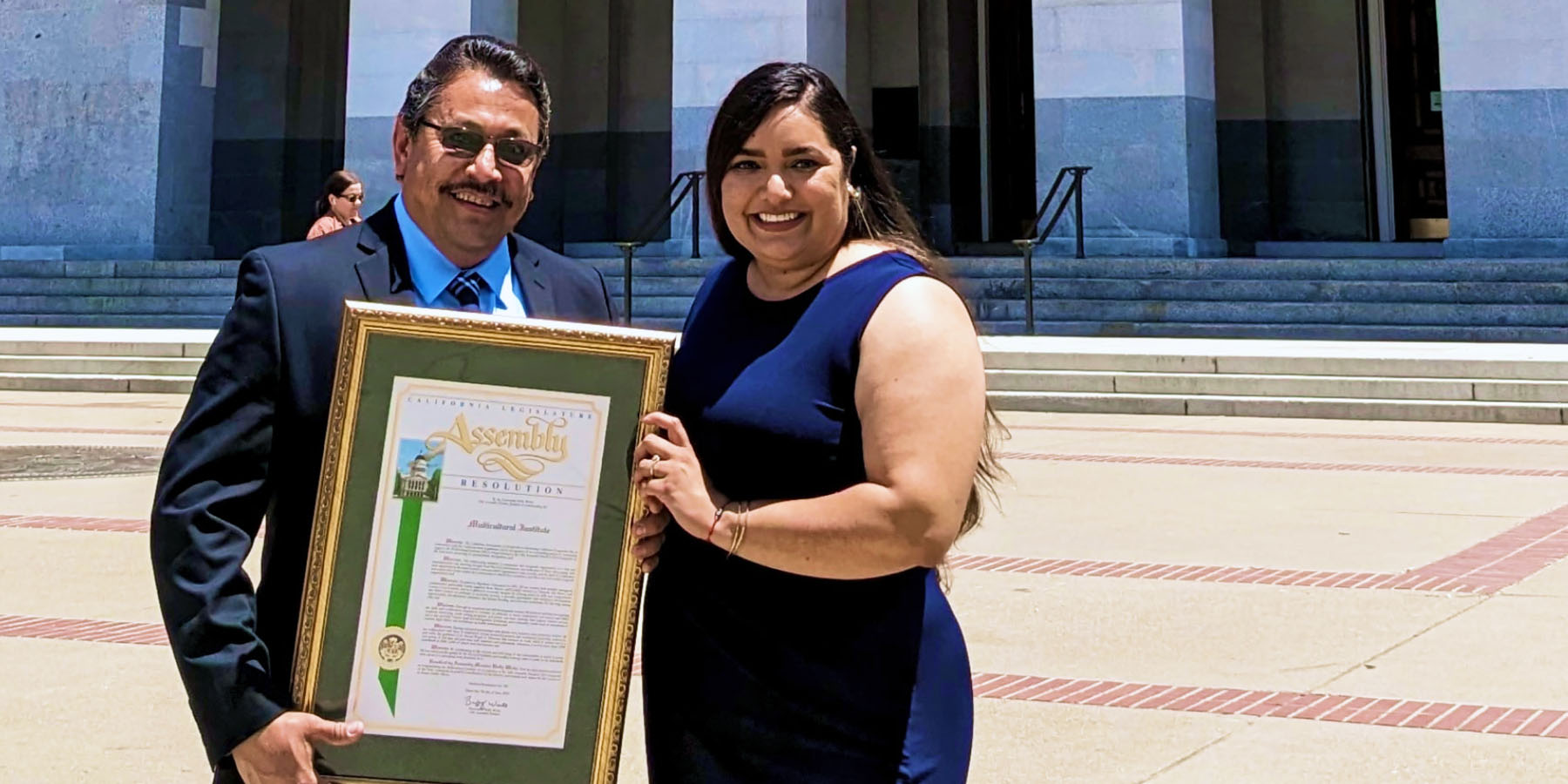 Mirna Cervantes smiles while standing beside a man holding a large framed declaration.