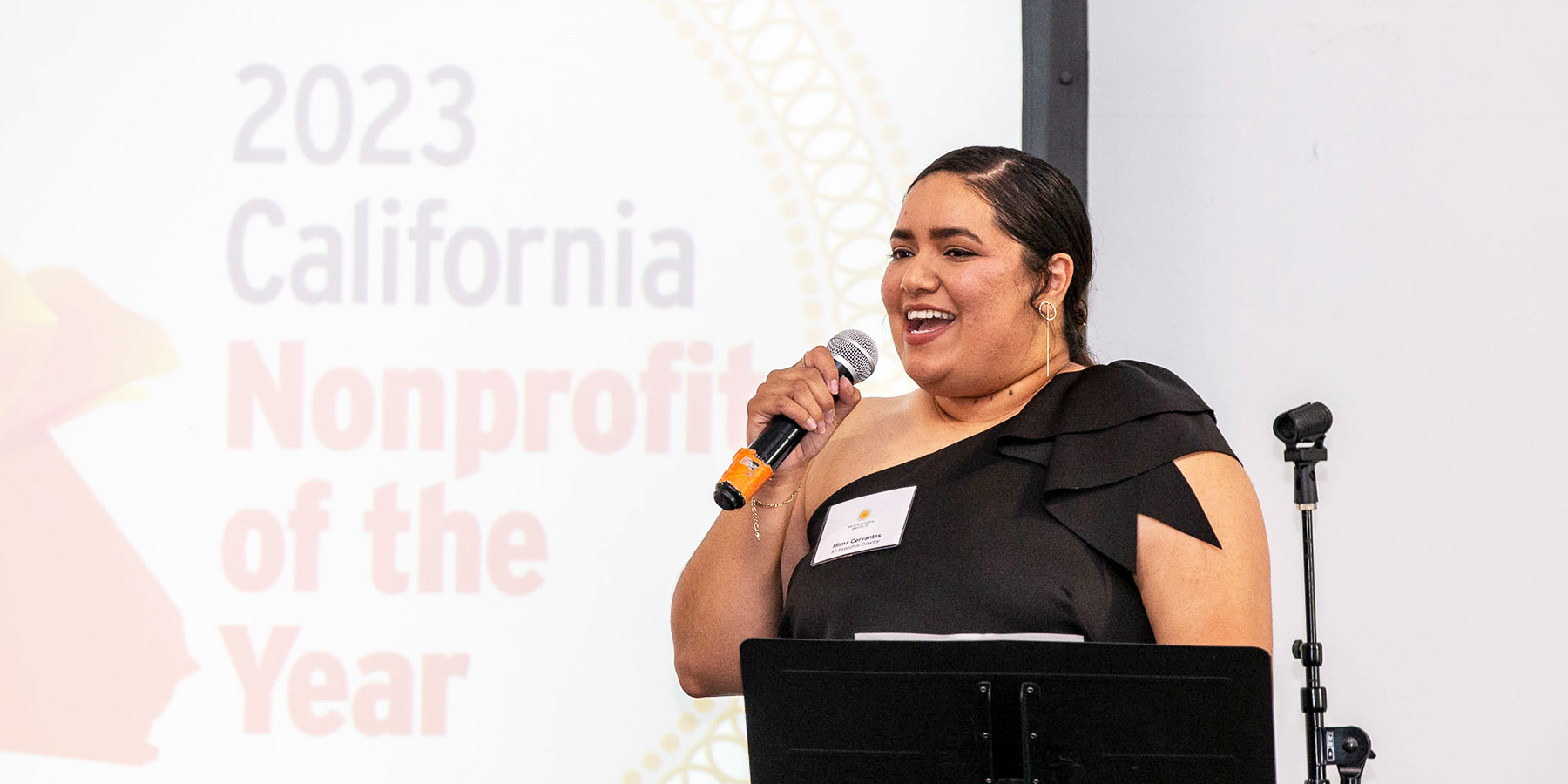 Mirna Cervantes stands behind a podium and speaks into a microphone. A screen behind her reads, “2023 California Nonprofit of the Year.”