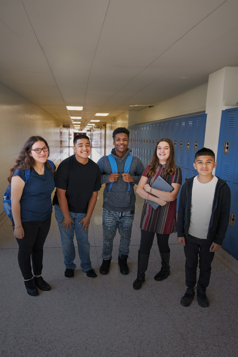 Five teenaged students stand in a school hallway surrounded by lockers.