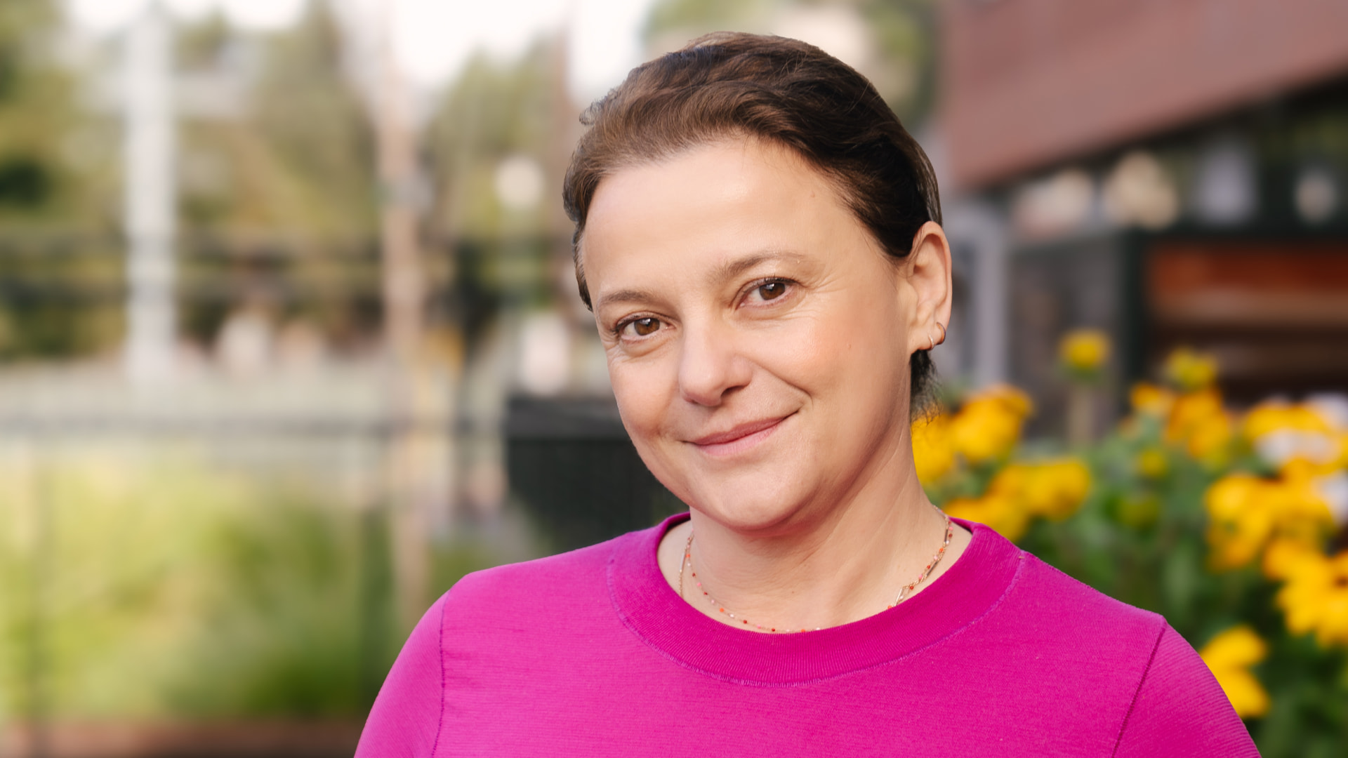 Dr. Raluca Kurz, wearing a hot pink sweater, looks directly at the camera and smiles; yellow flowers and CZI headquarters are in the background.