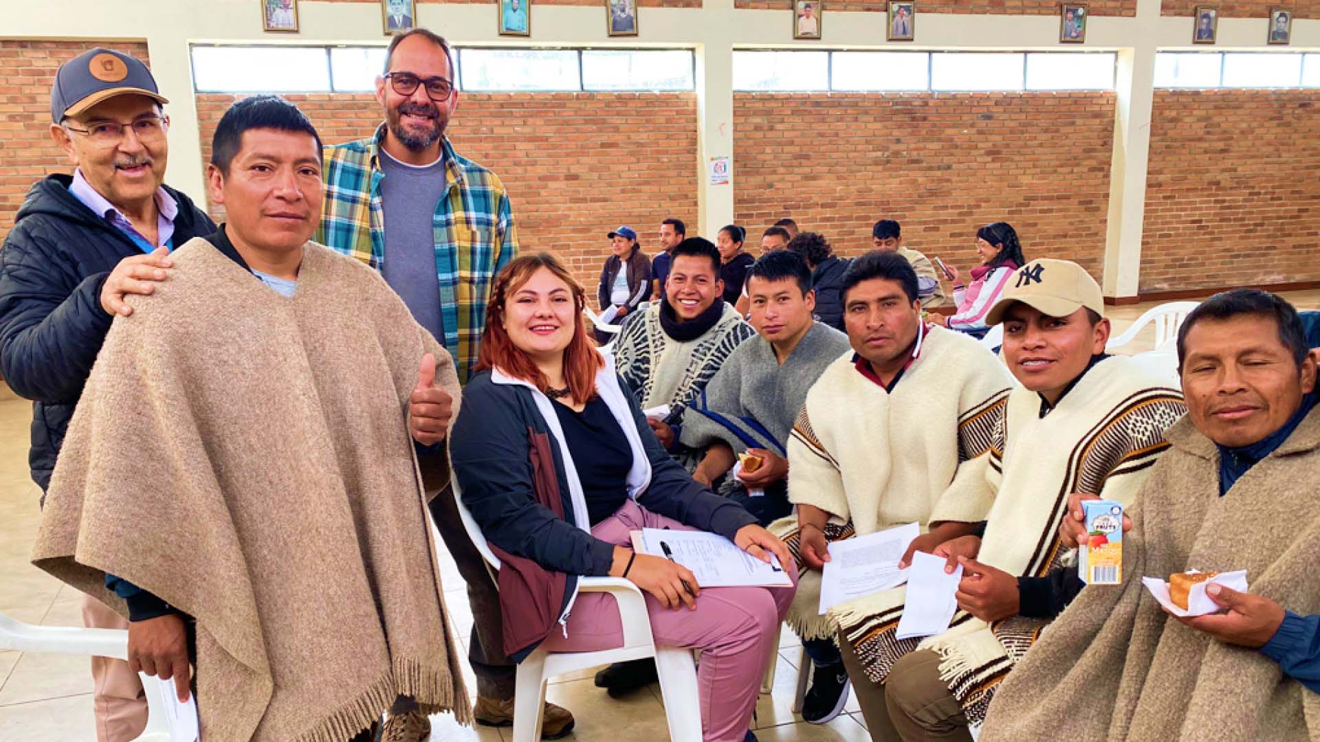 Members of the research team smiling with members of the community who are wearing cream, gray or light brown ponchos while sitting on white plastic chairs in a brick building