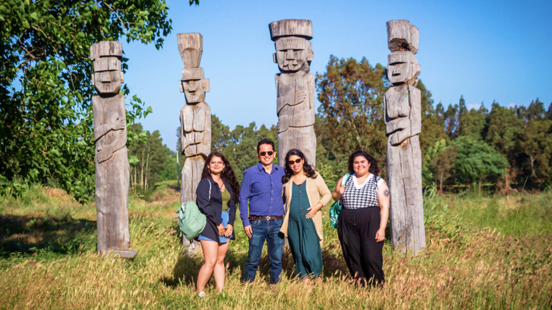 Four members of the Chilean research team in a grassy field with trees all around smiling while posing in front of four tall, stone-carved statues