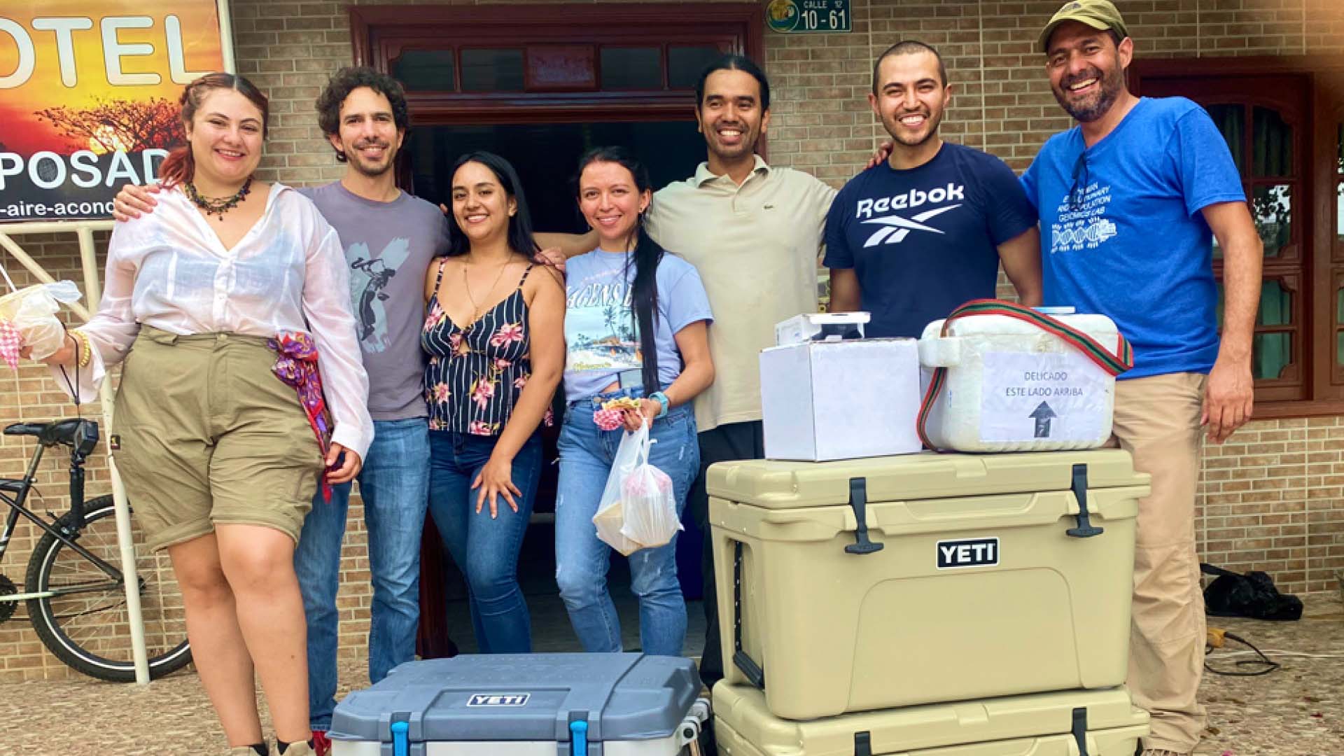 Seven members of the research team smiling outside of a building with three large coolers and two ice boxes stacked in front of them