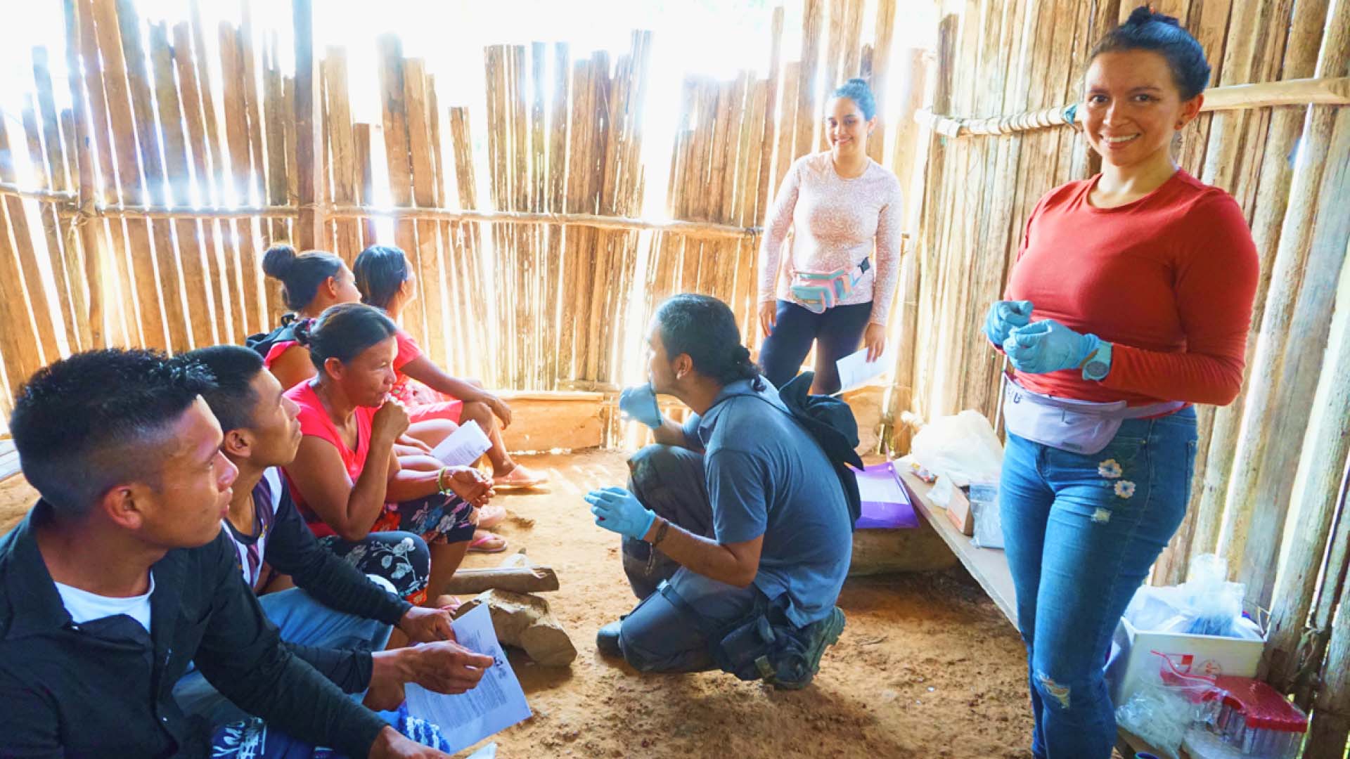 Local sampling coordinator Marisol Espitia stands next to two research team members, smiling, with one of them crouching while talking to members of the local community. The researchers are wearing blue gloves and are all gathered in a sunny room with vertical wood planks making up the walls