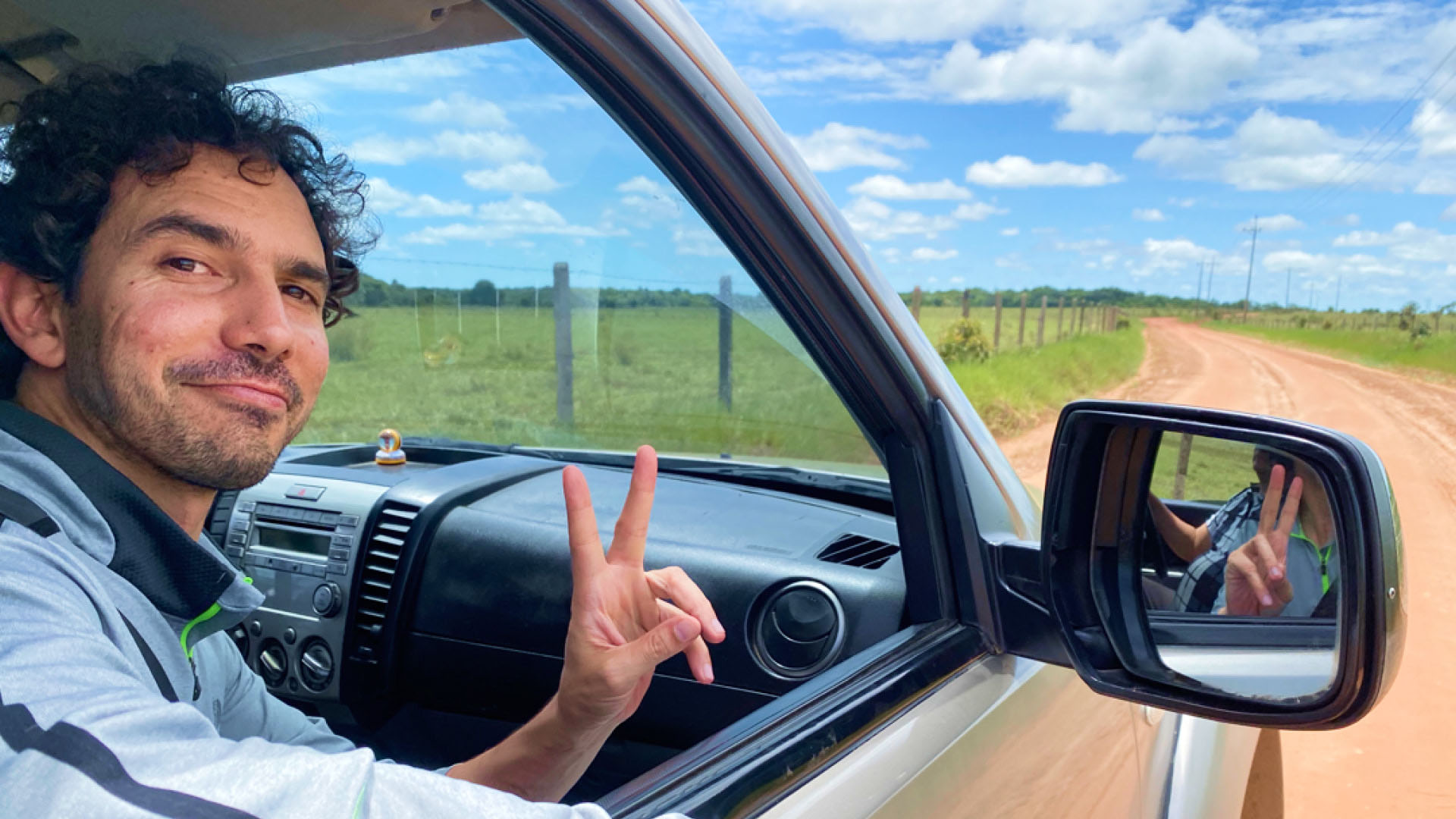 Carlos smirking out of the passenger side window of a silver car, holding his hands up in a peace sign