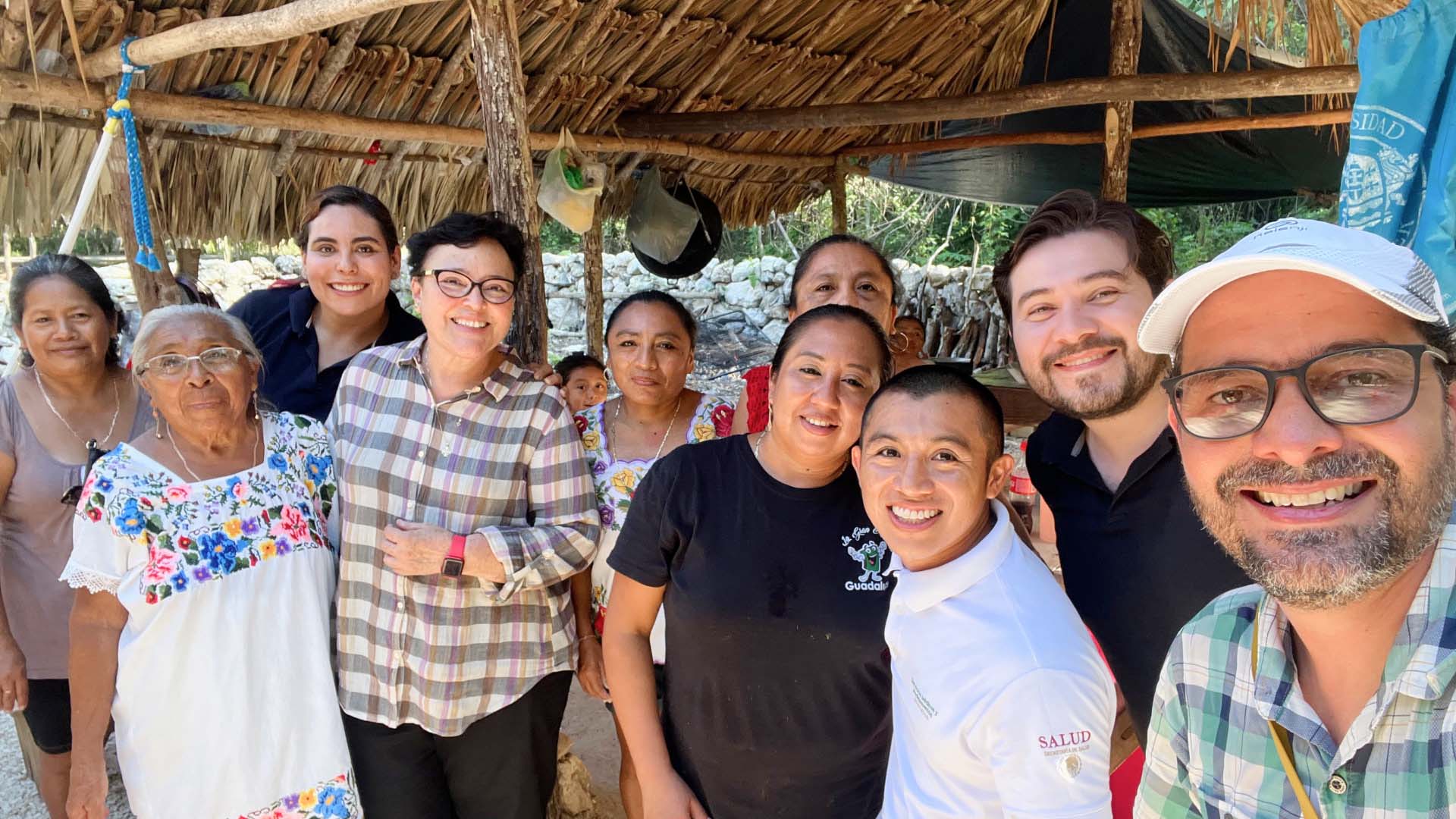 Selfie taken by Andrés, smiling in a cap and glasses in a green and blue checkered shirt with a group of people from the Mayan community smiling next to the research team, all huddled under a wooden-roofed structure.