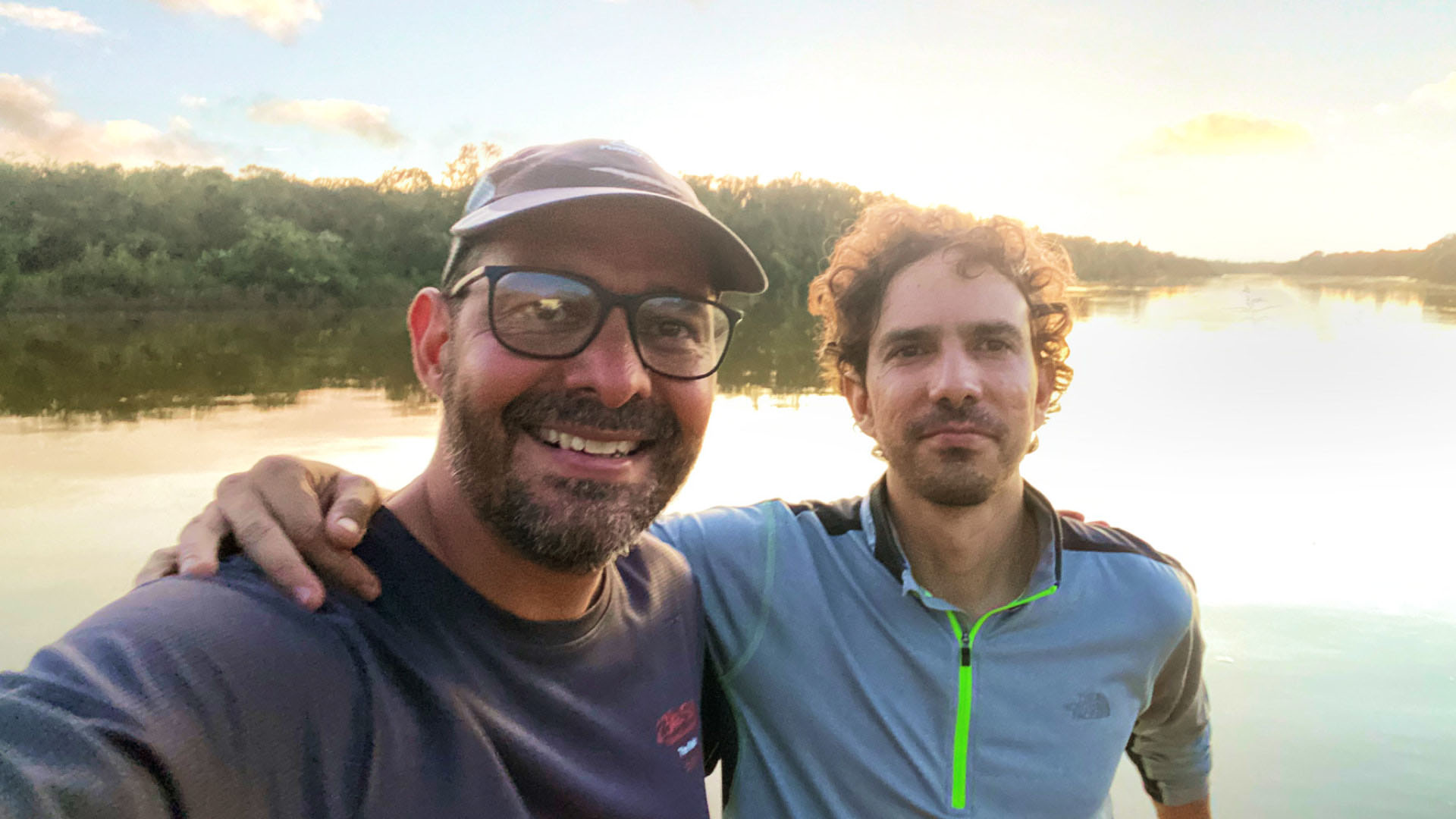Selfie in front of a sun-reflecting river with Andrés wearing a cap and glasses, smiling next to Carlos, who is leaning on the edge of the boat with an arm around Andrés