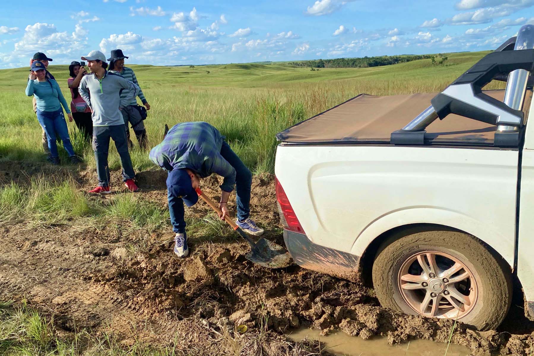 A group of people wait while a person in a baseball cap works to dig a white truck out of the mud.