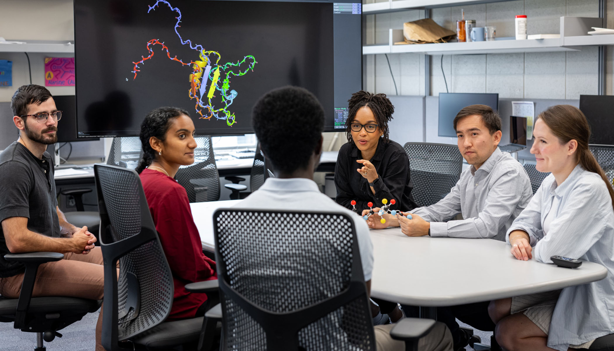 Group of researchers is sitting together at a table in a lab and talking with each other. In the background, a screen is shown depicting a photo of a protein.