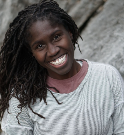 A Black Woman with dreadlocks is wearing a white striped shirt, smiling at the camera with an offset mountain backdrop.