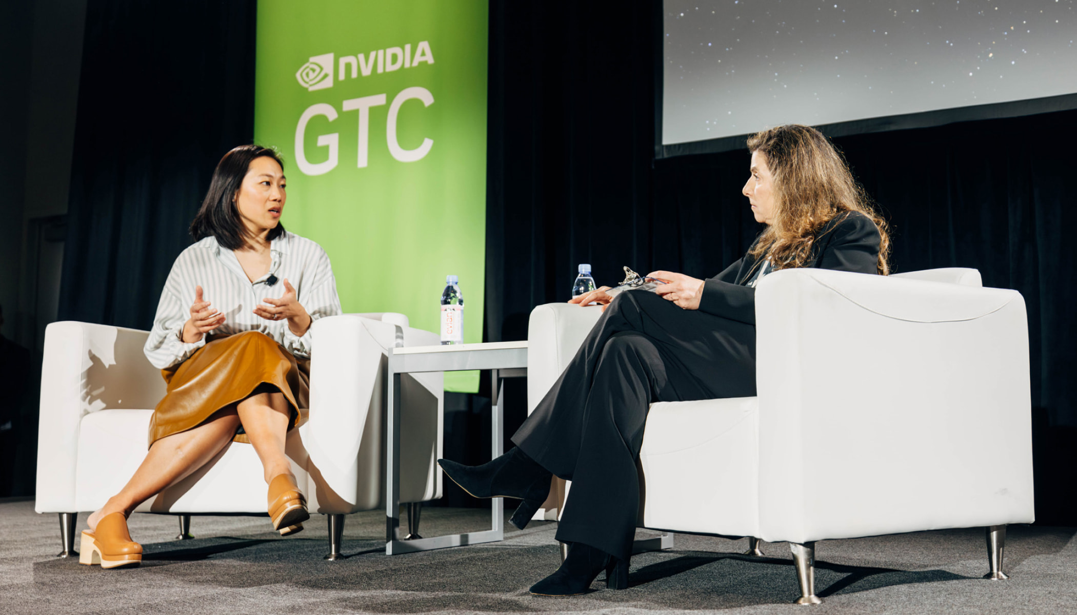 Drs. Priscilla Chan and Mona G. Flores sit in white armchairs on a stage for a fireside chat. A green banner that reads “NVIDIA GTC” hangs behind them.