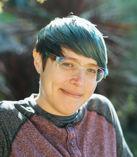 A photo of Ananda Valenzuela, a light-skinned non-binary human with short brown hair, glasses, and a big smile.