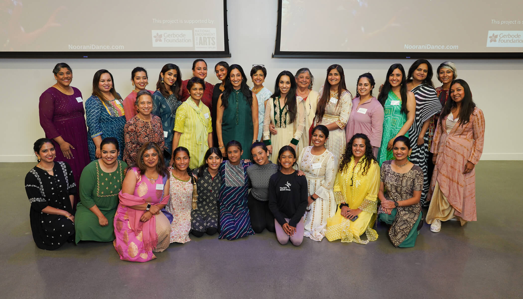 A large group of women in colorful traditional South Asian attire pose for a photo in front of two large screens.