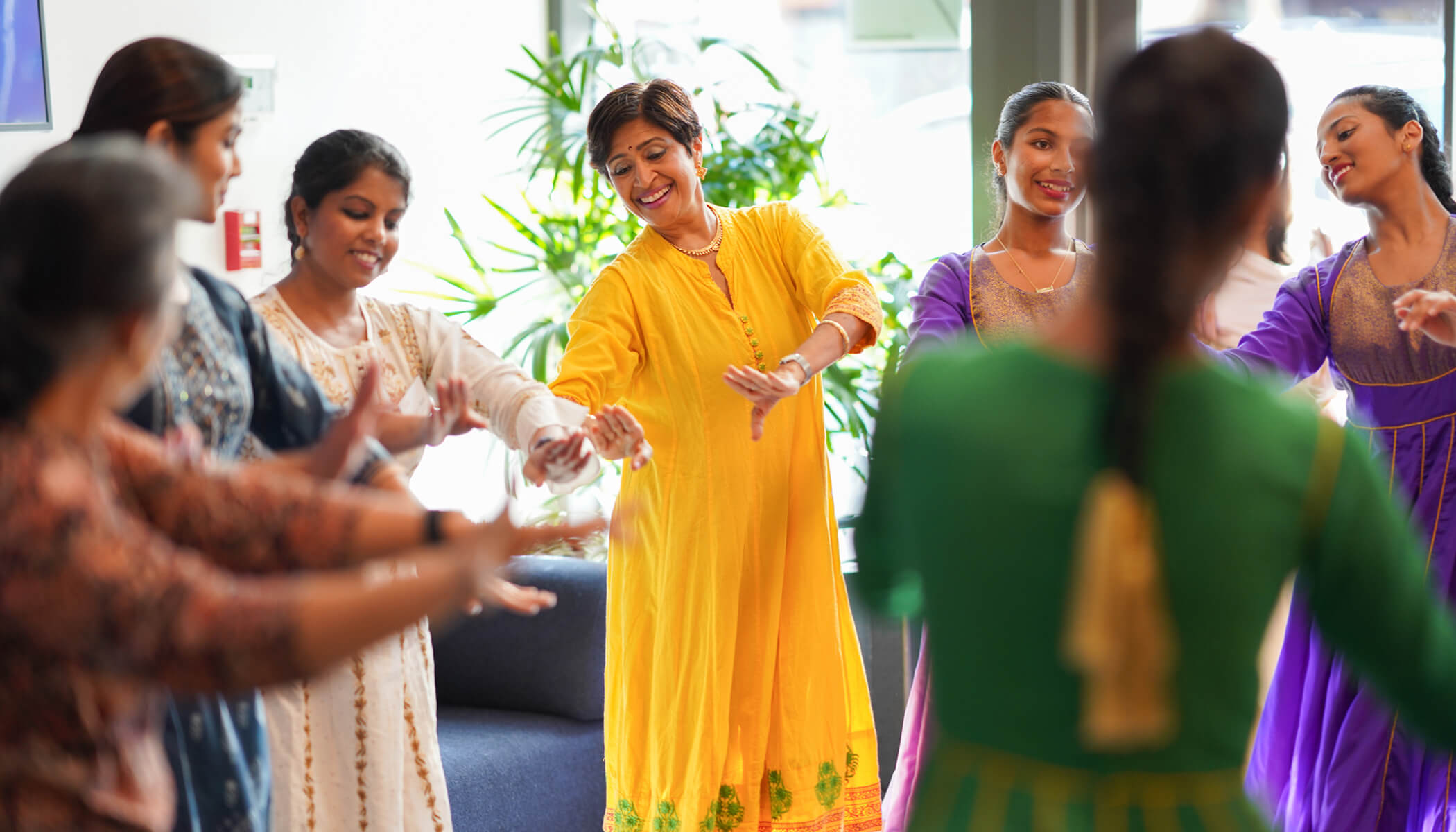 A group of women in colorful traditional South Asian attire are smiling and dancing together with hand gestures in an indoor setting with plants in the background.