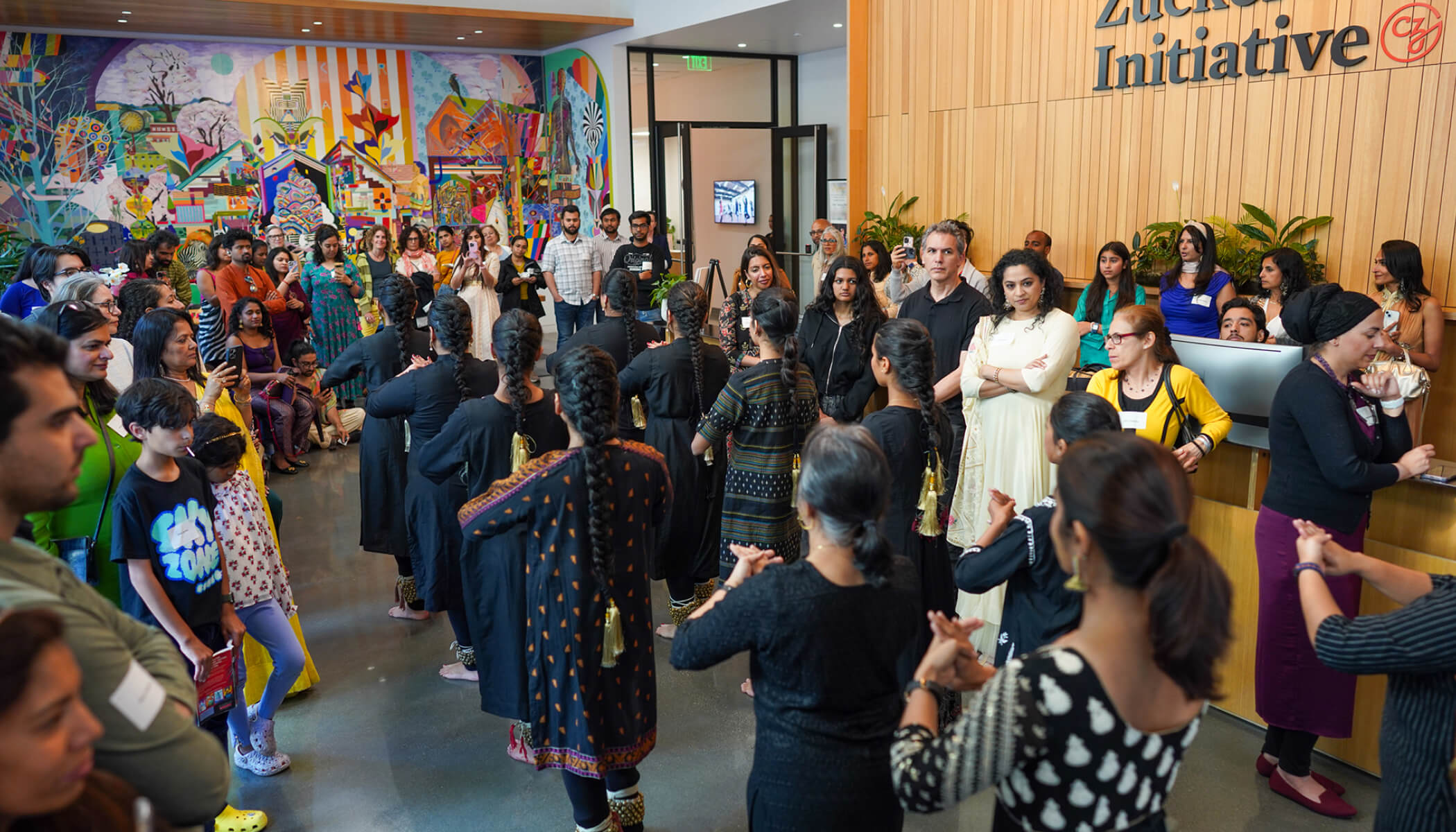 A group of women in Black, traditional South Asian attire dance in unison through a lobby. “Chan Zuckerberg Initiative” is on the wall.