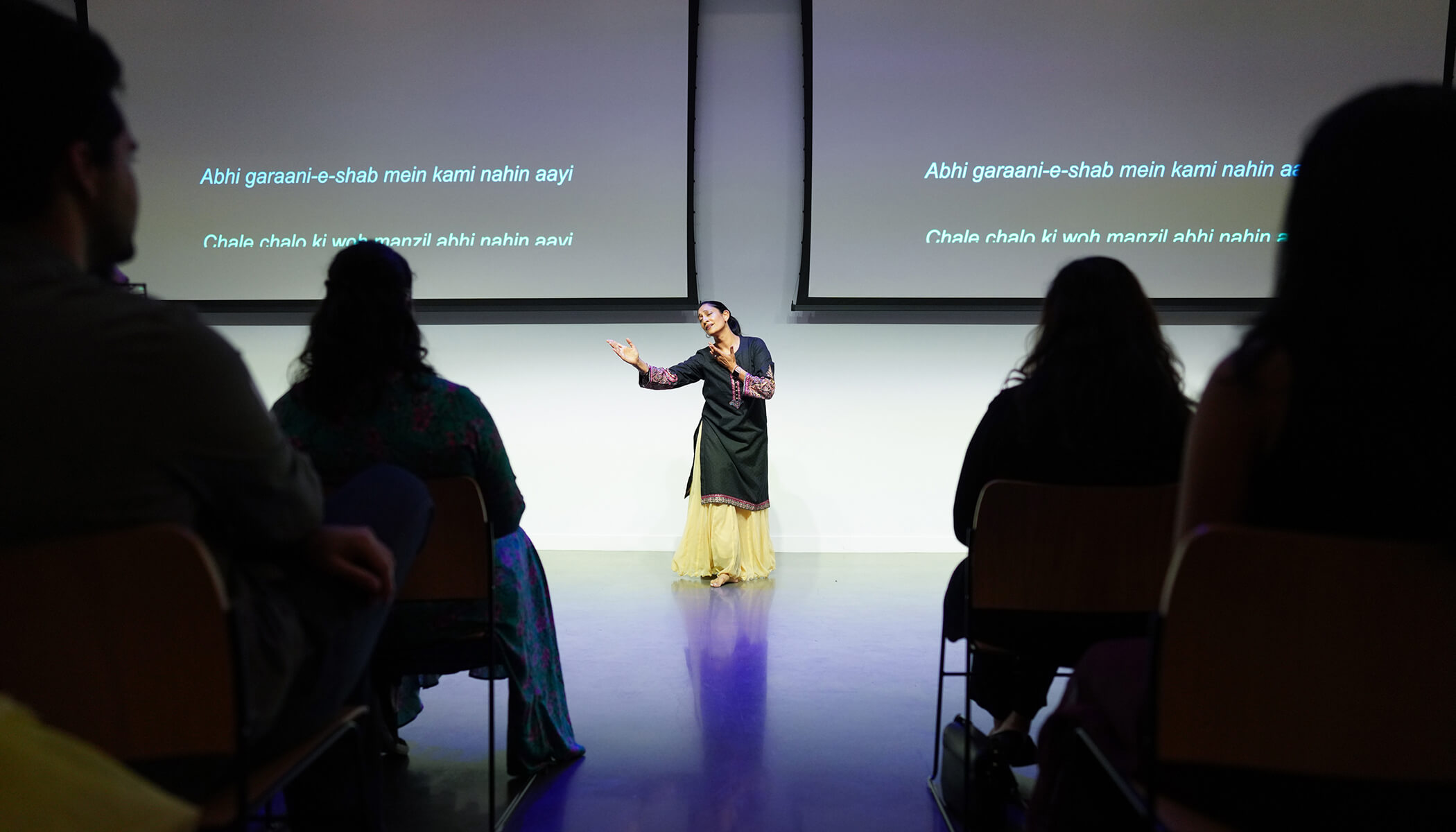 A woman in black and yellow dances in front of two screens with Urdu words on them. Shadows of audience members are in the foreground.