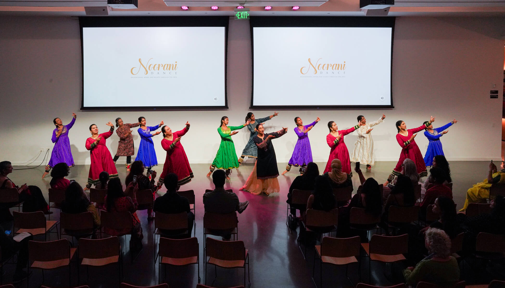 A group of women in colorful South Asian attire dance in unison in front of two screens that read “Noorani.”