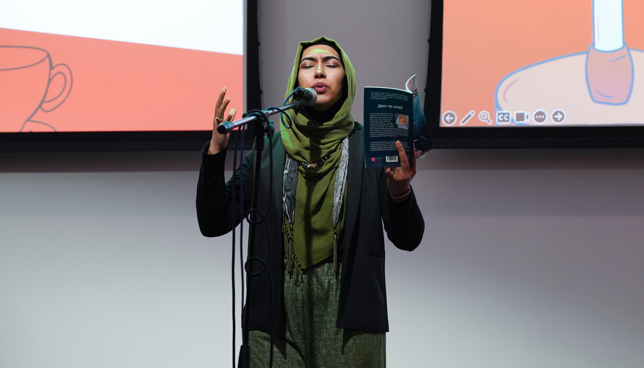 A woman in a green hijab reads from a book before a mic. Her hand is raised in expression.