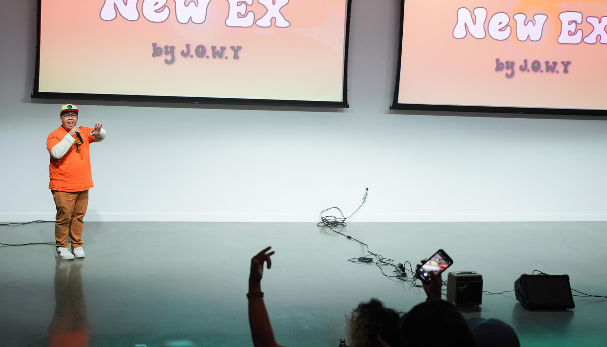 A person in an orange t-shirt and baseball cap talking into a microphone in front of an audience. Screens that say “New Ex by J.O.W.Y” are in the background.
