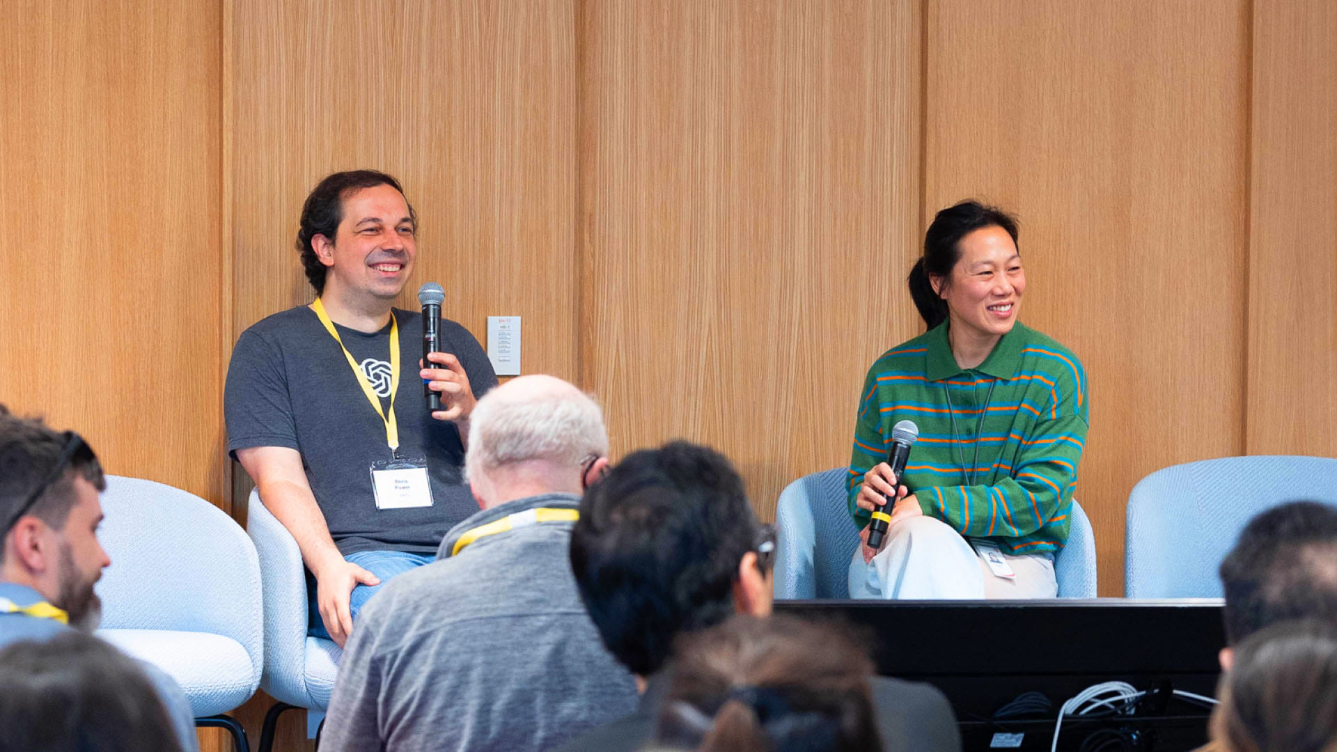Two people are seated on a panel, both holding microphones and smiling; the man on the left, Boris Power, is wearing a T-shirt with a logo and a lanyard, while the woman on the right, Priscilla Chan, is wearing a green striped shirt. An audience is visible in the foreground.