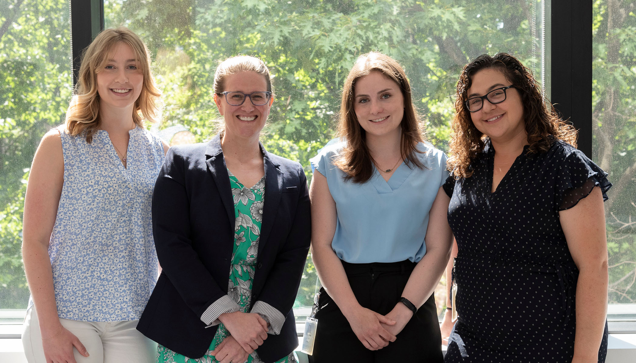 Katie Russell, Samantha Baxter, Carmen Glaze, and Moriel Singer-Berk stand together smiling in front of a window with green foliage in the background, representing the team from the Broad Institute's Center for Mendelian Genomics involved in creating the GeniE tool.