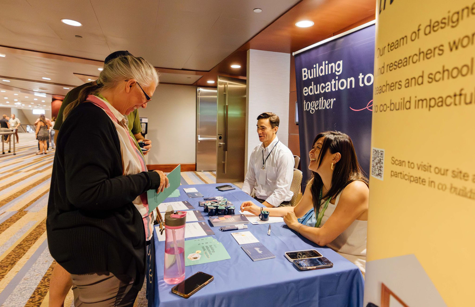 A woman standing behind a table in front of a Render Exhibit booth interacting with a man and women seated behind the table