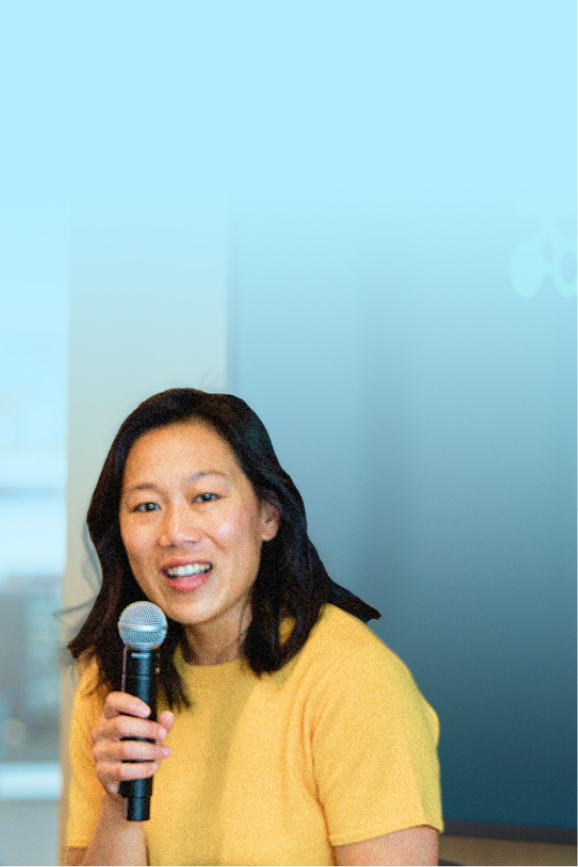 Priscilla Chan is speaking into a microphone, smiling, and set against a light blue background.