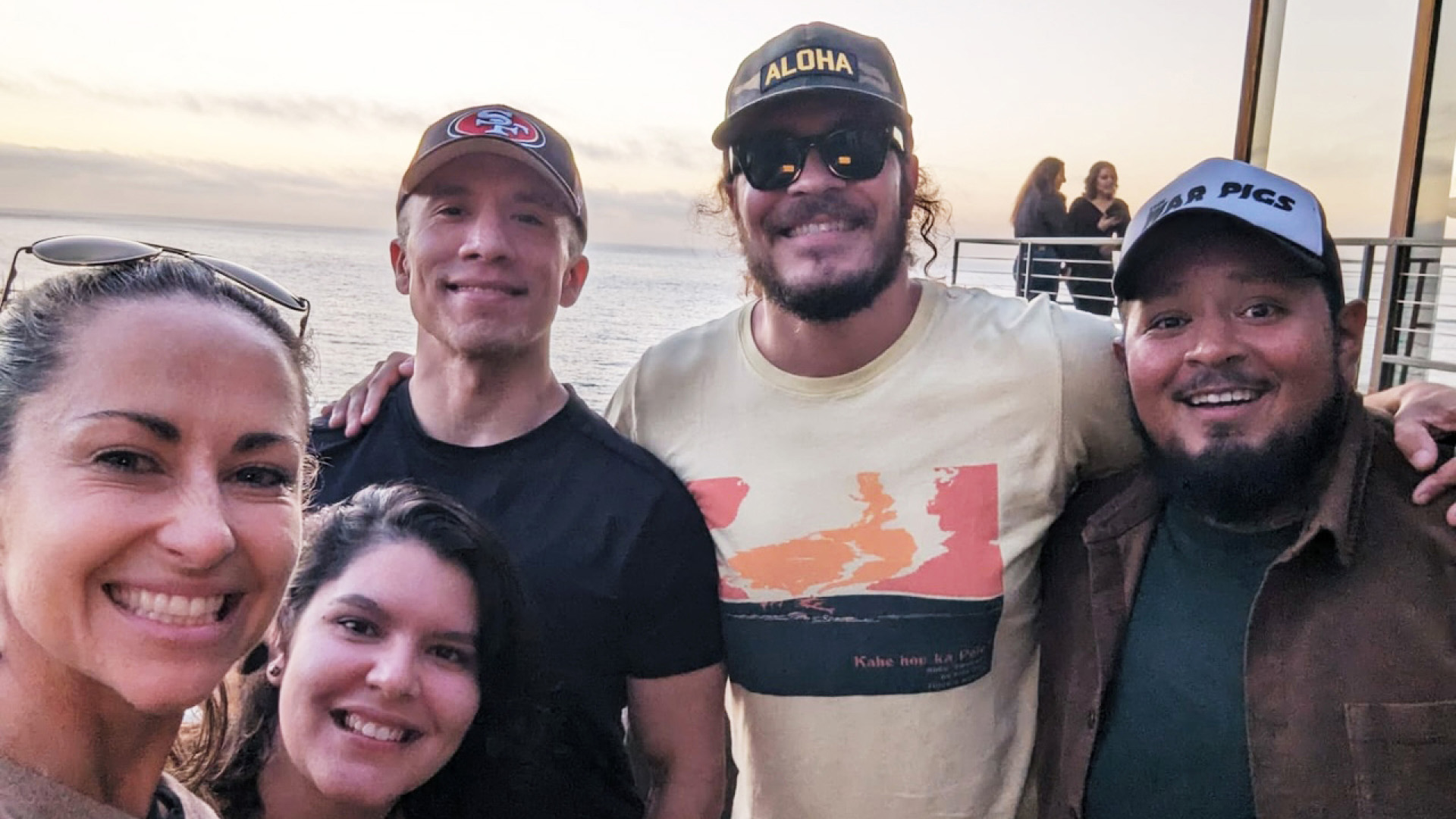 Five people smile at the camera with an ocean sunset in the background.