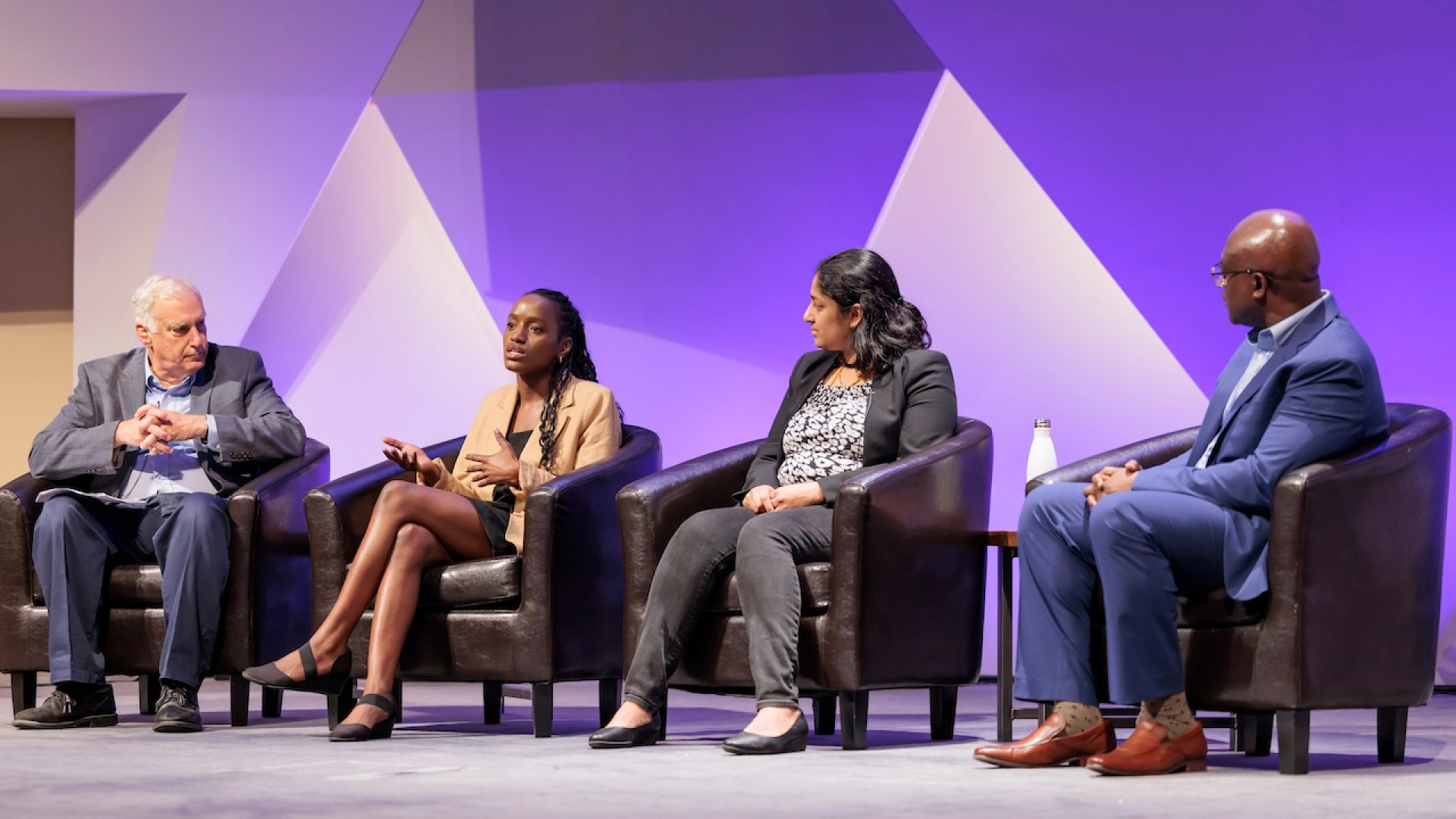 Four panelists sit on stage in armchairs, engaged in a discussion, with one person speaking while the others listen attentively.