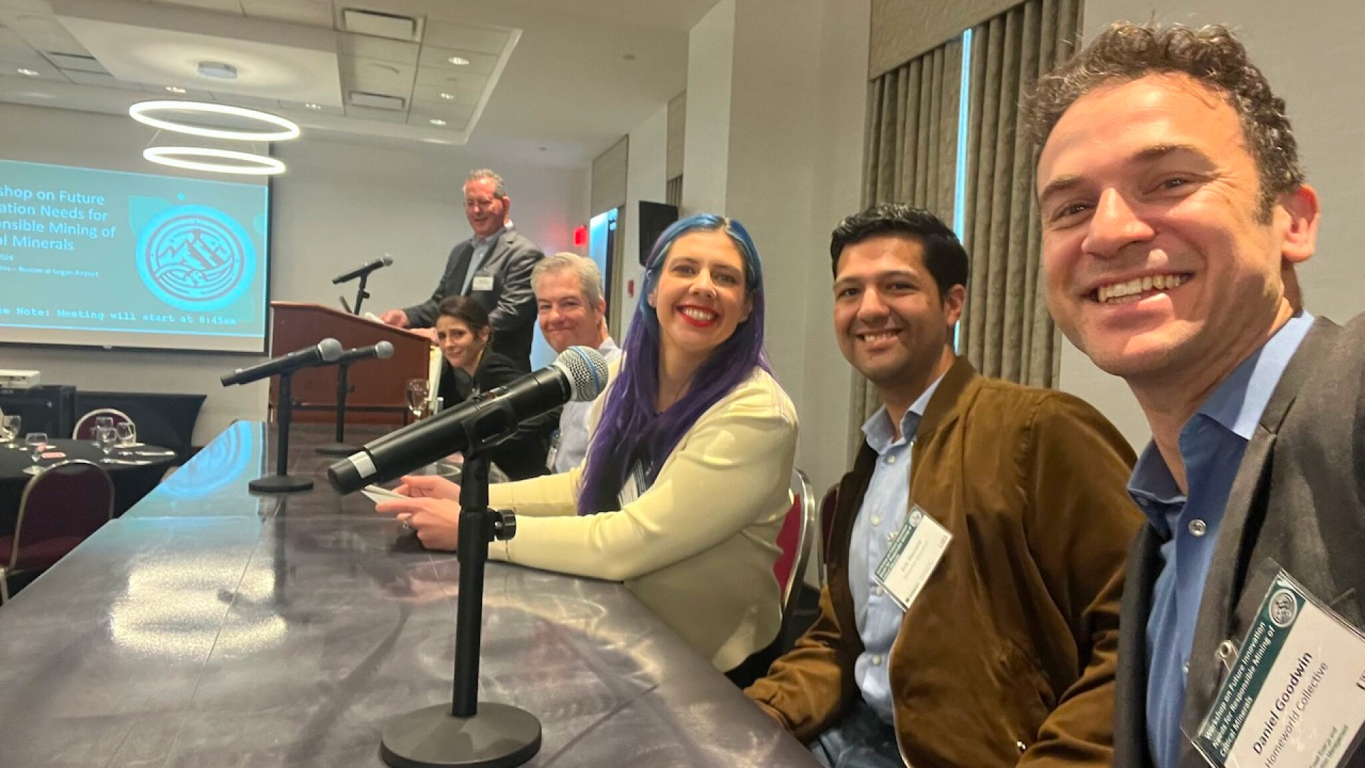 A group of five panelists sits at a table, smiling, with a presentation on the responsible mining of critical minerals displayed behind them.