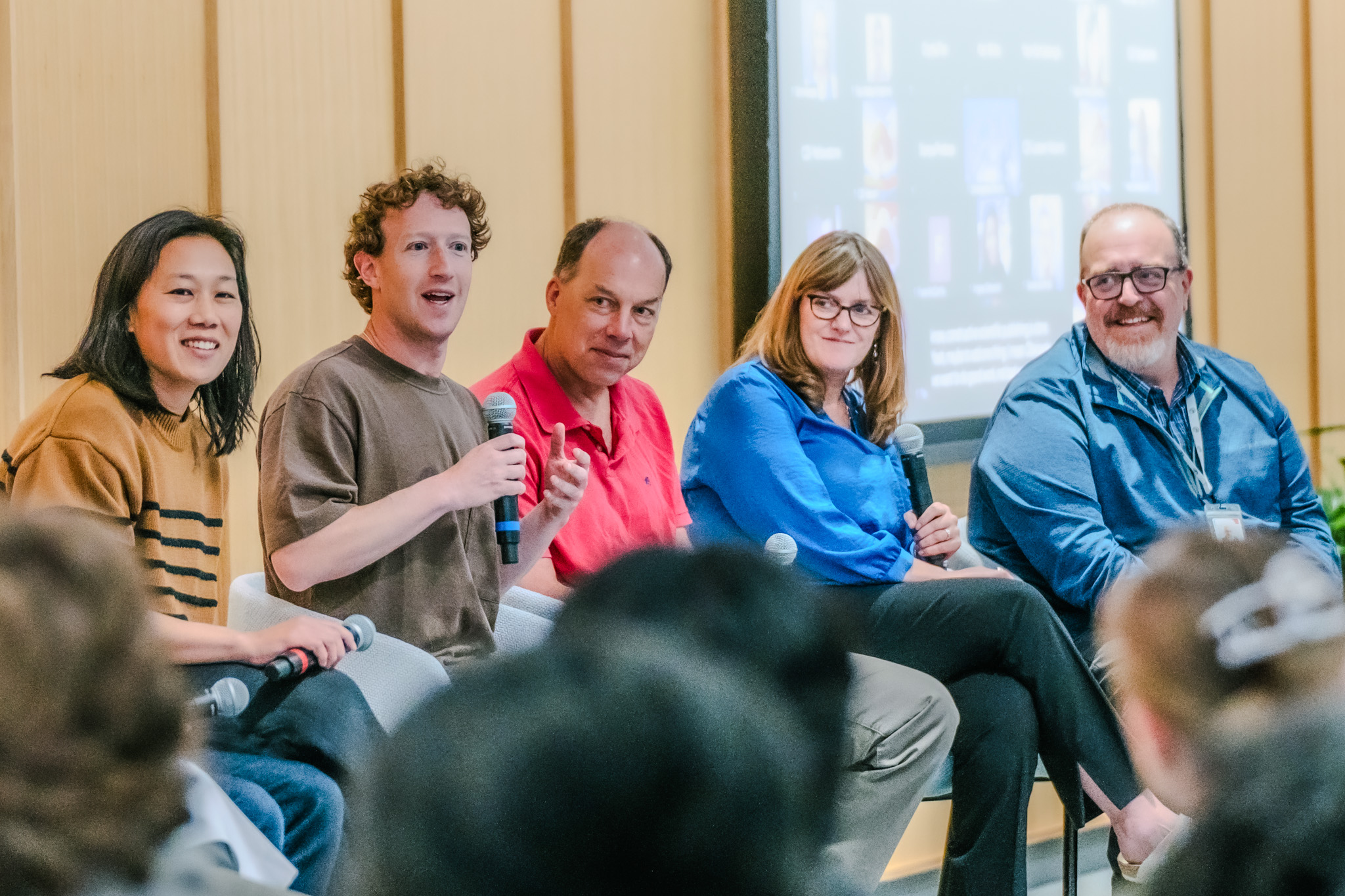 A panel of five people seated and holding microphones, engaged in a discussion; Mark Zuckerberg is speaking while the others listen attentively and smile.