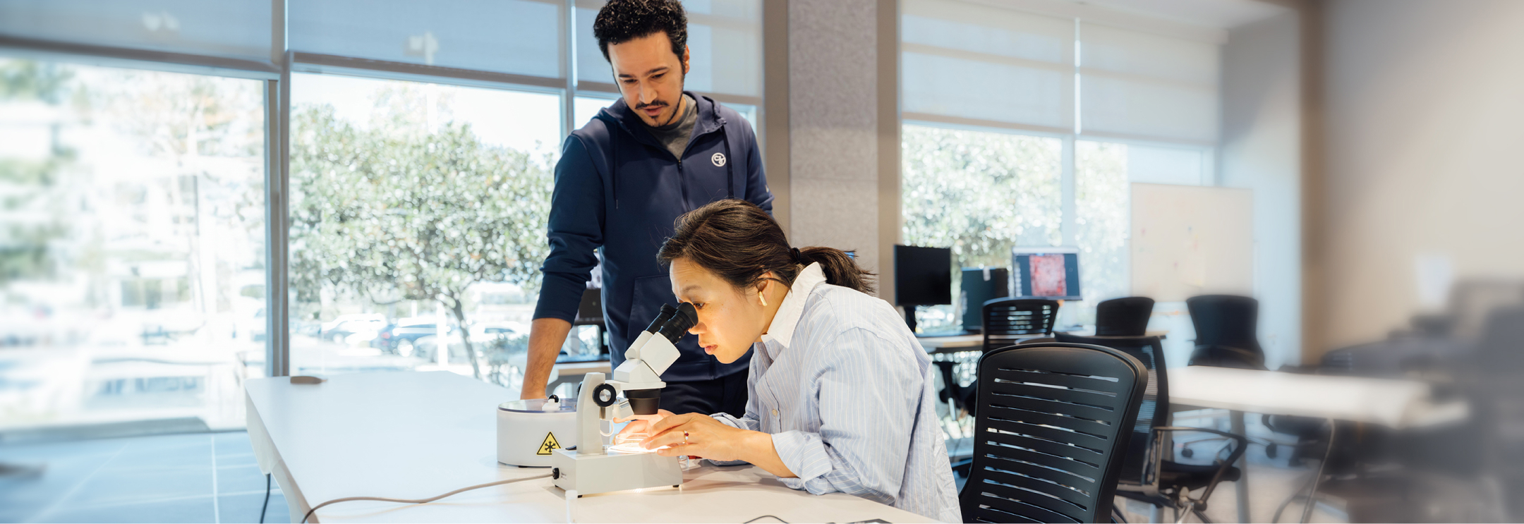 Priscilla Chan is using a microscope in a well-lit lab, focusing on a specimen while a colleague stands beside her, observing.