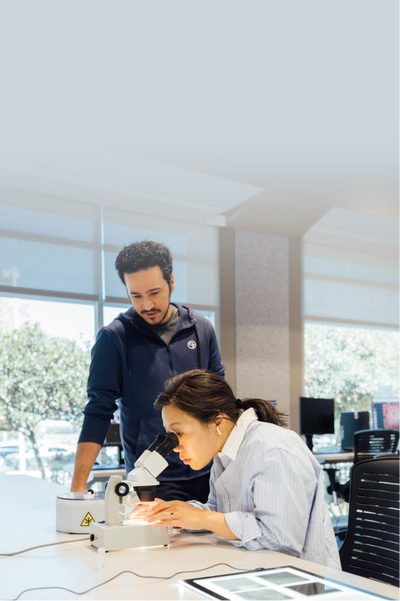 Priscilla Chan is focused on looking through a microscope while a colleague observes her work in a brightly lit laboratory, set against a light blue background.