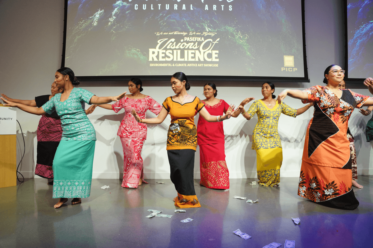 A group of dancers dressed in colorful garb raise their arms at their sides in unison as they perform a traditional Pacific Islander dance.