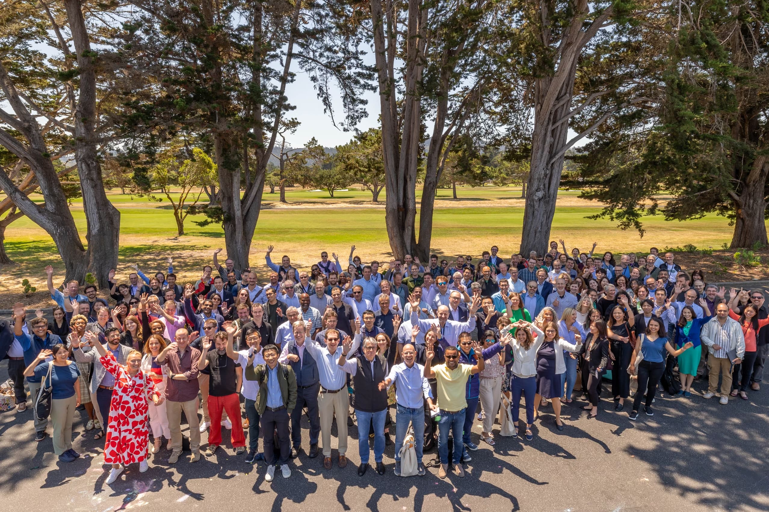 Participants of the Neuroscience 2024 Meeting stand and look up to the camera waving and smiling. They stand under trees outside on a sunny day.