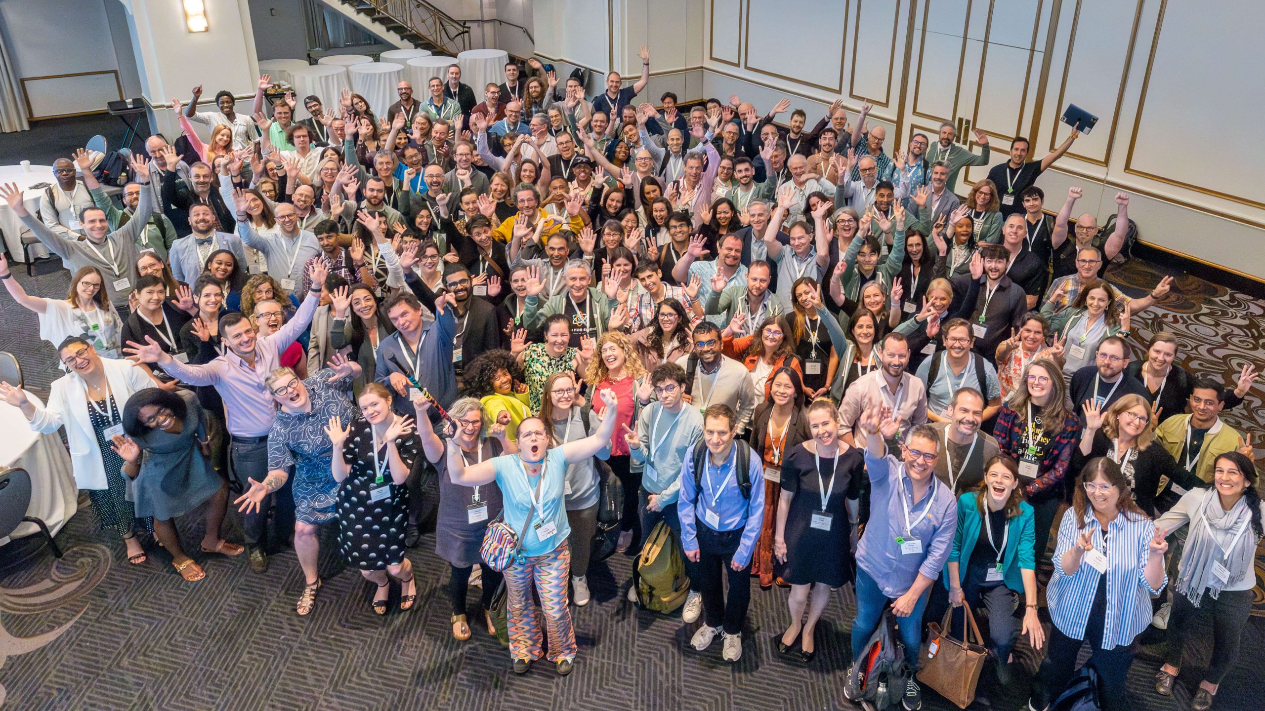 A group of Open Science 2024 Meeting participants look up toward the camera and enthusiastically wave and smile.