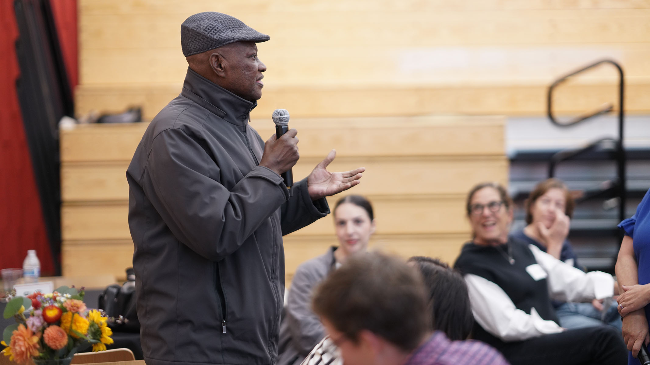 A man in a dark gray hat and jacket speaks into a microphone and uses hand gestures as attendees listen.