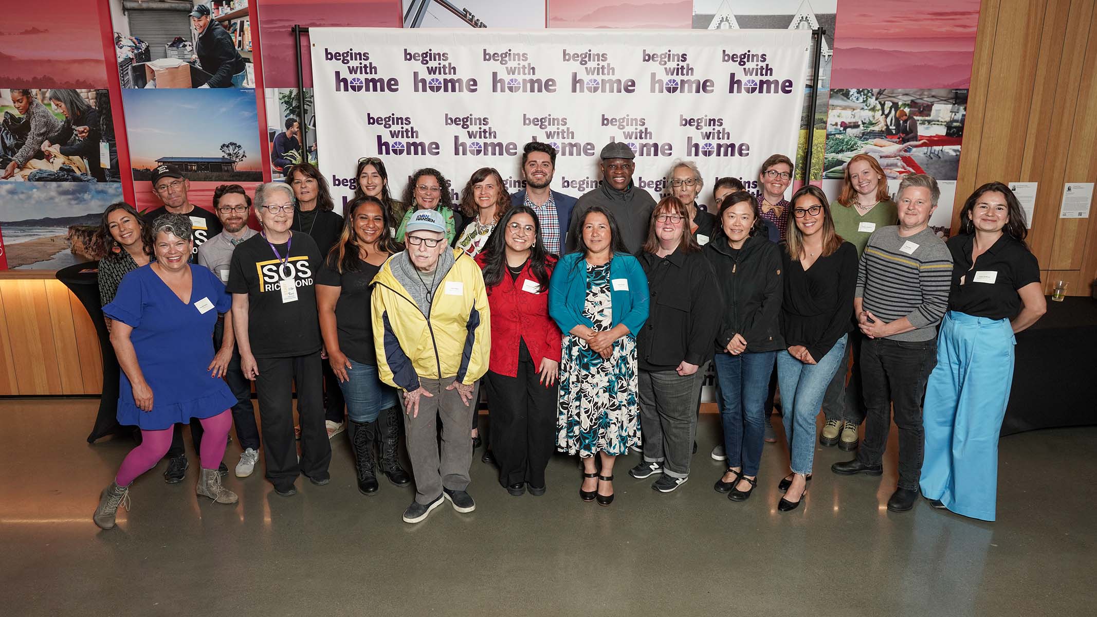 About two dozen people stand together and smile in front of a backdrop that says, “Begins with Home.”