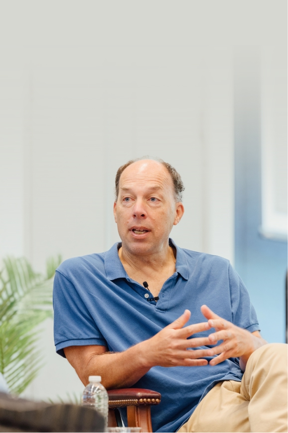 Steve Quake is speaking, gesturing with his hands during a discussion, wearing a blue polo shirt, with a calm expression.