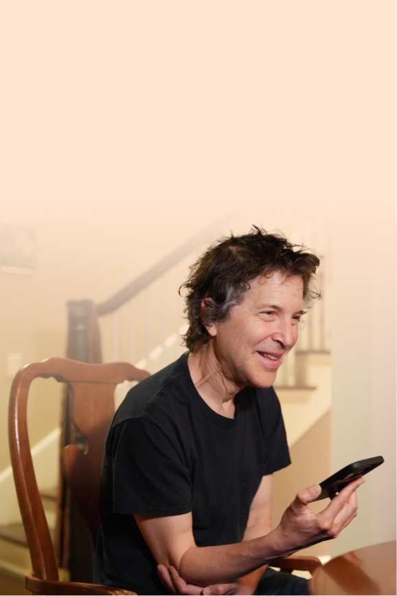 David Baker in a black t-shirt and jeans, sitting at a dining table smiling with his cell phone in his hand.