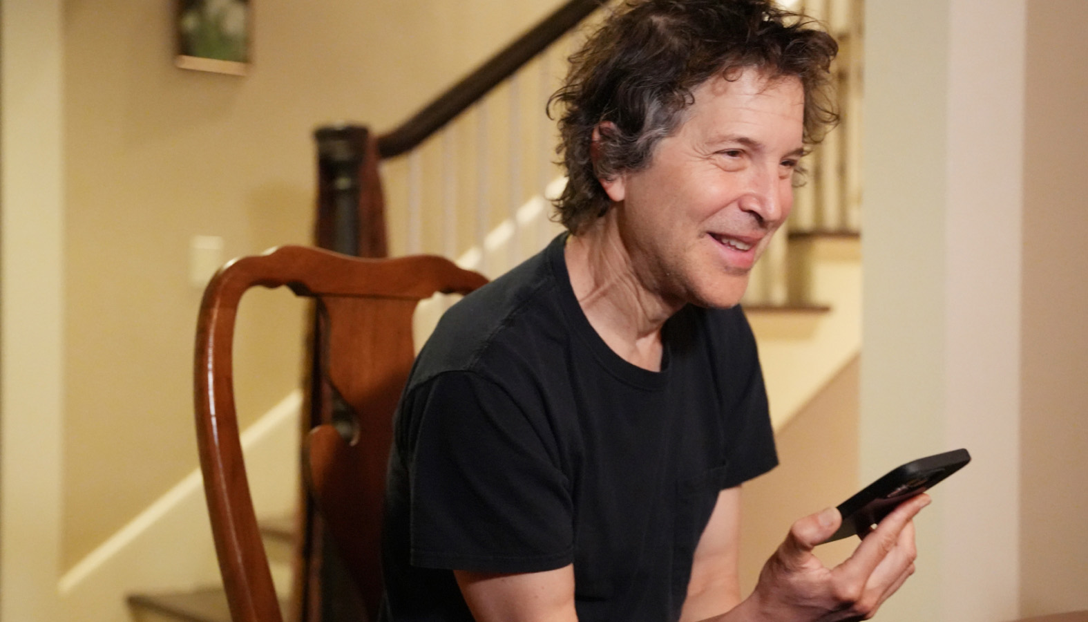 David Baker in a black t-shirt, sitting in a dining chair, smiling with his cell phone in his hand.