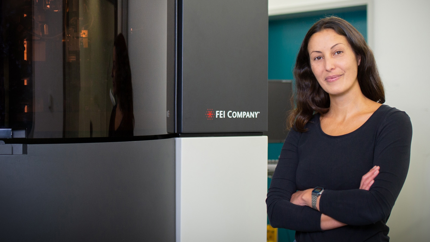 Julia Mahamid in a dark, long-sleeved shirt with arms crossed, smiling in front of a large box structure containing a microscope.