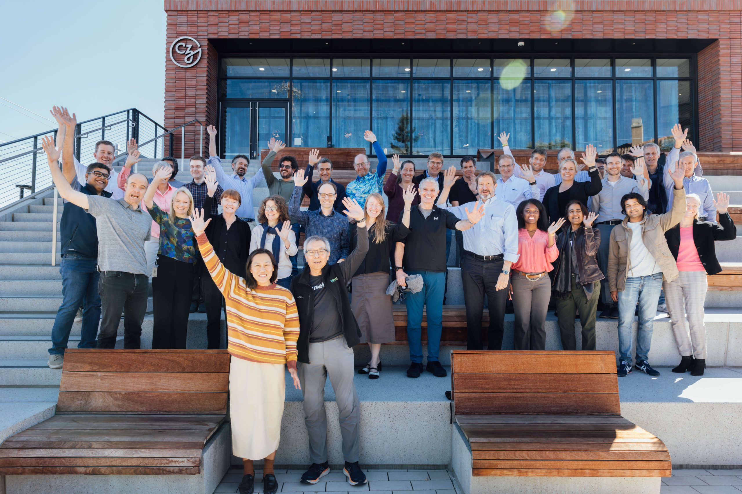 Meeting participants gather close together to smile and wave at the camera. They pose together outside of the CZI office on wooden and cement benches.