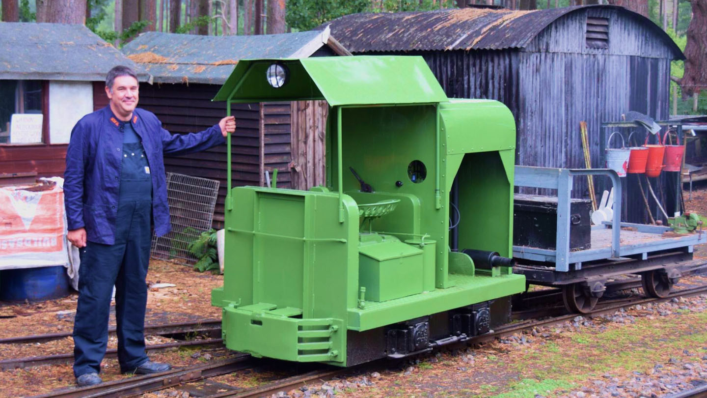 Dan Quine, in a blue work outfit, stands next to a small green locomotive on narrow-gauge tracks, with rustic sheds and tall trees in the background.