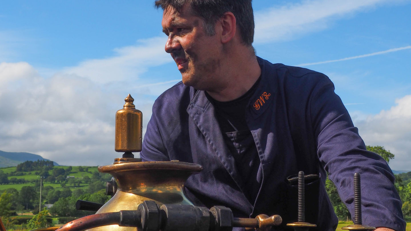 Dan Quine, wearing a blue work jacket with “GWR” embroidered on the collar, smiles while sitting beside a brass steam engine component.